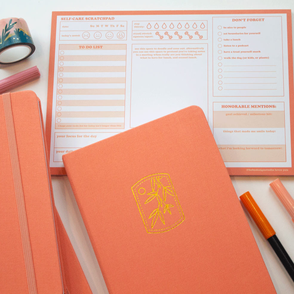 Three tangerine bobo BuJo dot grid journals arranged with other orange desk supplies. The gold stamp illustration and elastic closure can be seen on the journals.