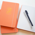 two tangerine bobo journals stacked on top of each other and an open one next to the stack with two pens on top