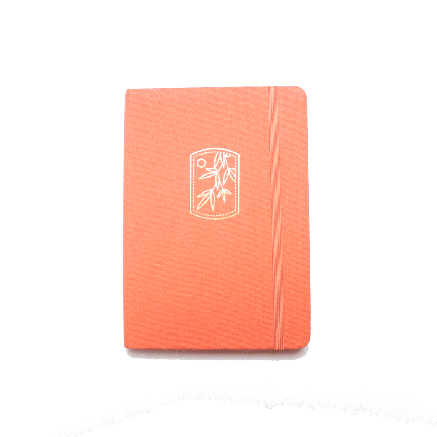 The A5 bobo BuJo Dot Grid Journal in "Tangerine" orange. A gold foil illustration of mandarin tree leaves and a sun is stamped on the front cover. A matching orange elastic closure is holding the journal closed.