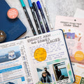 a blue open wanderlust passport filled out with writing and photos. to the right of the passport is the desert road trip sticker book and two sticker sheets. above the open passport is a closed blue passport with two washi tapes on top and next to that are five various pens that come in the bundle