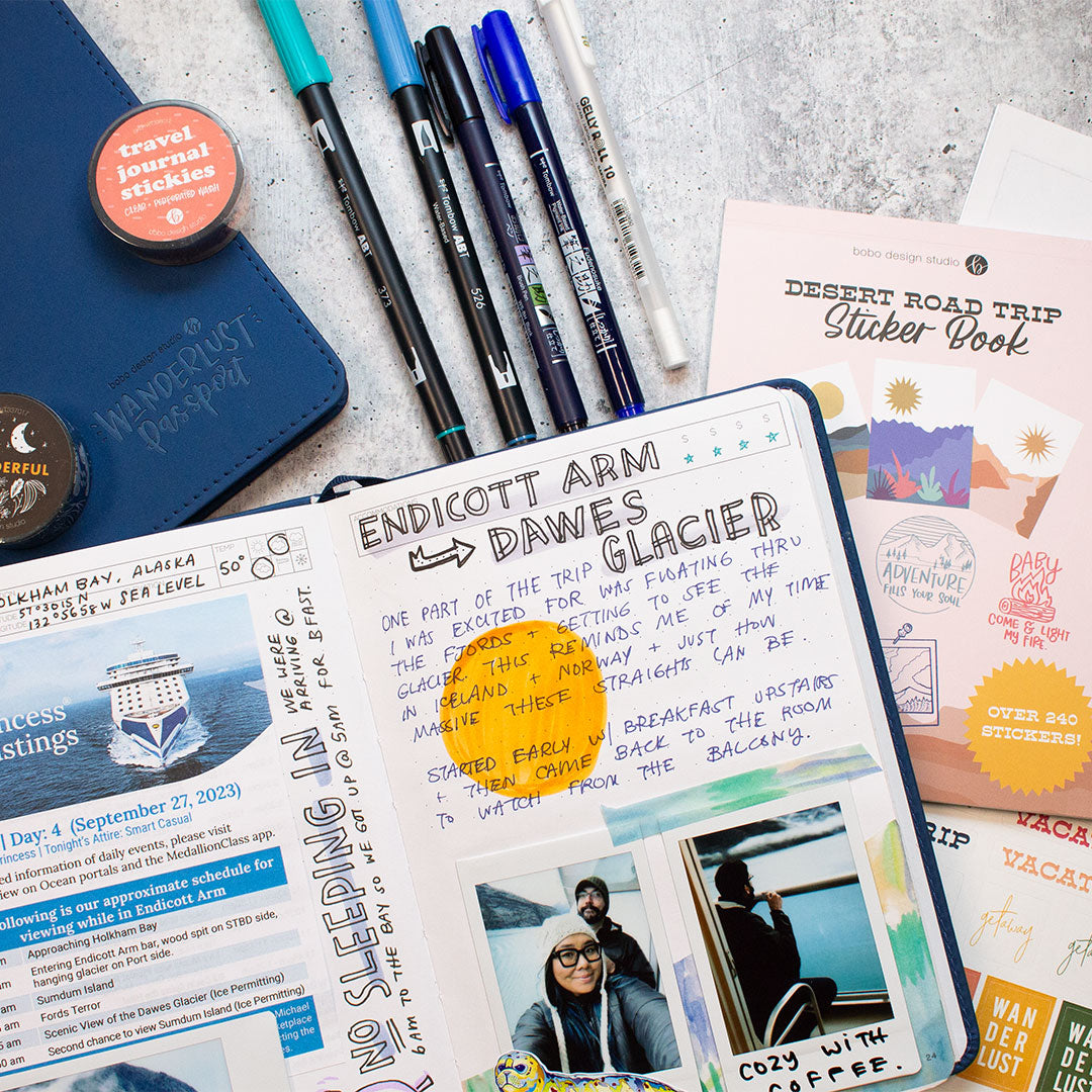 a blue open wanderlust passport filled out with writing and photos. to the right of the passport is the desert road trip sticker book and two sticker sheets. above the open passport is a closed blue passport with two washi tapes on top and next to that are five various pens that come in the bundle