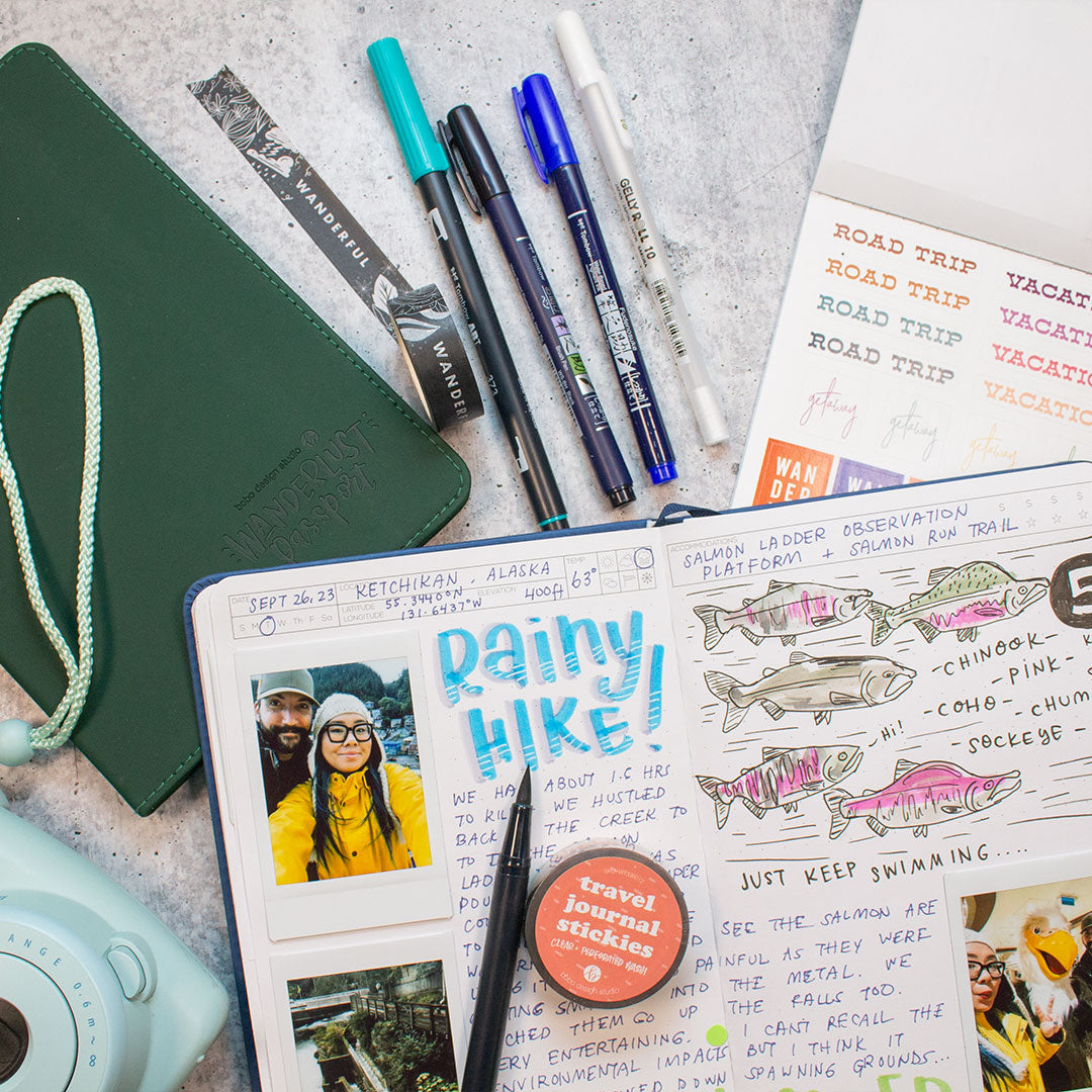 blue open wanderlust passport filled out with text, drawings, and photos. On top of book is an open brush pen and washi tape. next to the open passport is a light blue polaroid and a closed green wanderlust passport. above the open blue passport is a black open washi tape, four pens, and an open sticker sheet