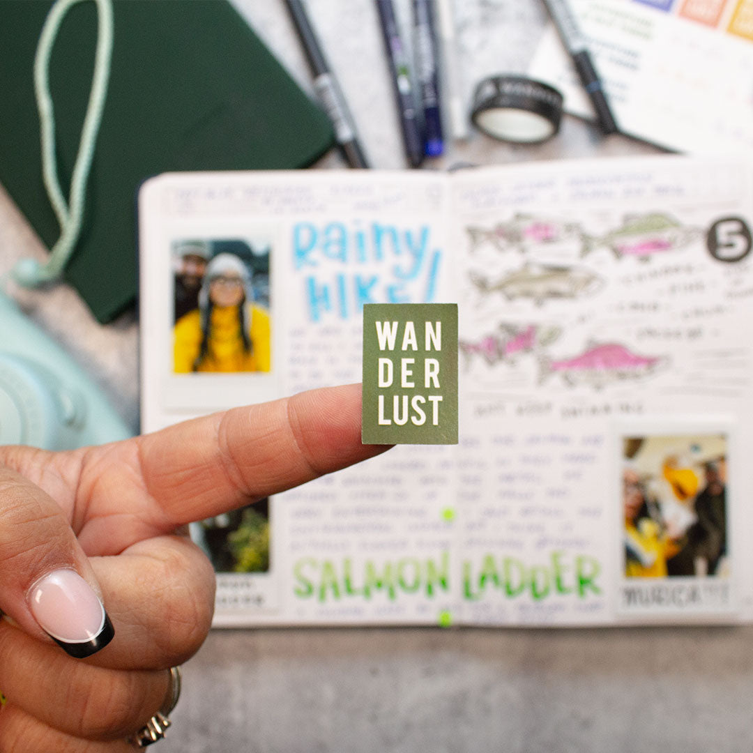 a left hand pointing a finger with a green sticker on the end of the finger that says wanderlust. underneath the hand is a filled out open wanderlust passport, a blue polaroid camera, a closed green wanderlust passport, five pens and a washi tape. 