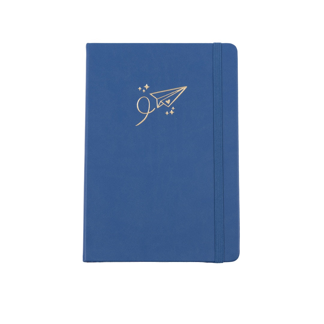 The A5 bobo Black dot grid journal in "airplane" with a medium blue vegan leather cover. A gold foil paper airplane with a heart and stars is stamped onto the front cover. A blue elastic closure is holding the journal shut.