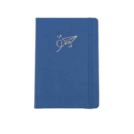 The A5 bobo Black dot grid journal in "airplane" with a medium blue vegan leather cover. A gold foil paper airplane with a heart and stars is stamped onto the front cover. A blue elastic closure is holding the journal shut.