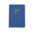 The A5 bobo Black dot grid journal in "airplane" with a medium blue vegan leather cover. A gold foil paper airplane with a heart and stars is stamped onto the front cover. A blue elastic closure is holding the journal shut.