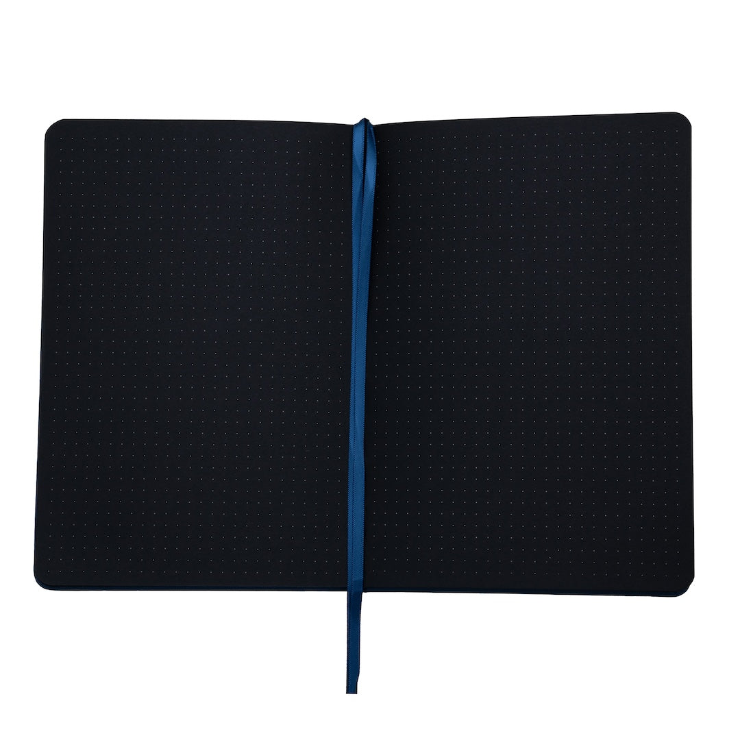 The bobo Black Dot Grid Journal is opened to show the black pages inside. The black A5 pages have small white dots arranged in a grid. The blue ribbon bookmark runs down the center of the pages spread.