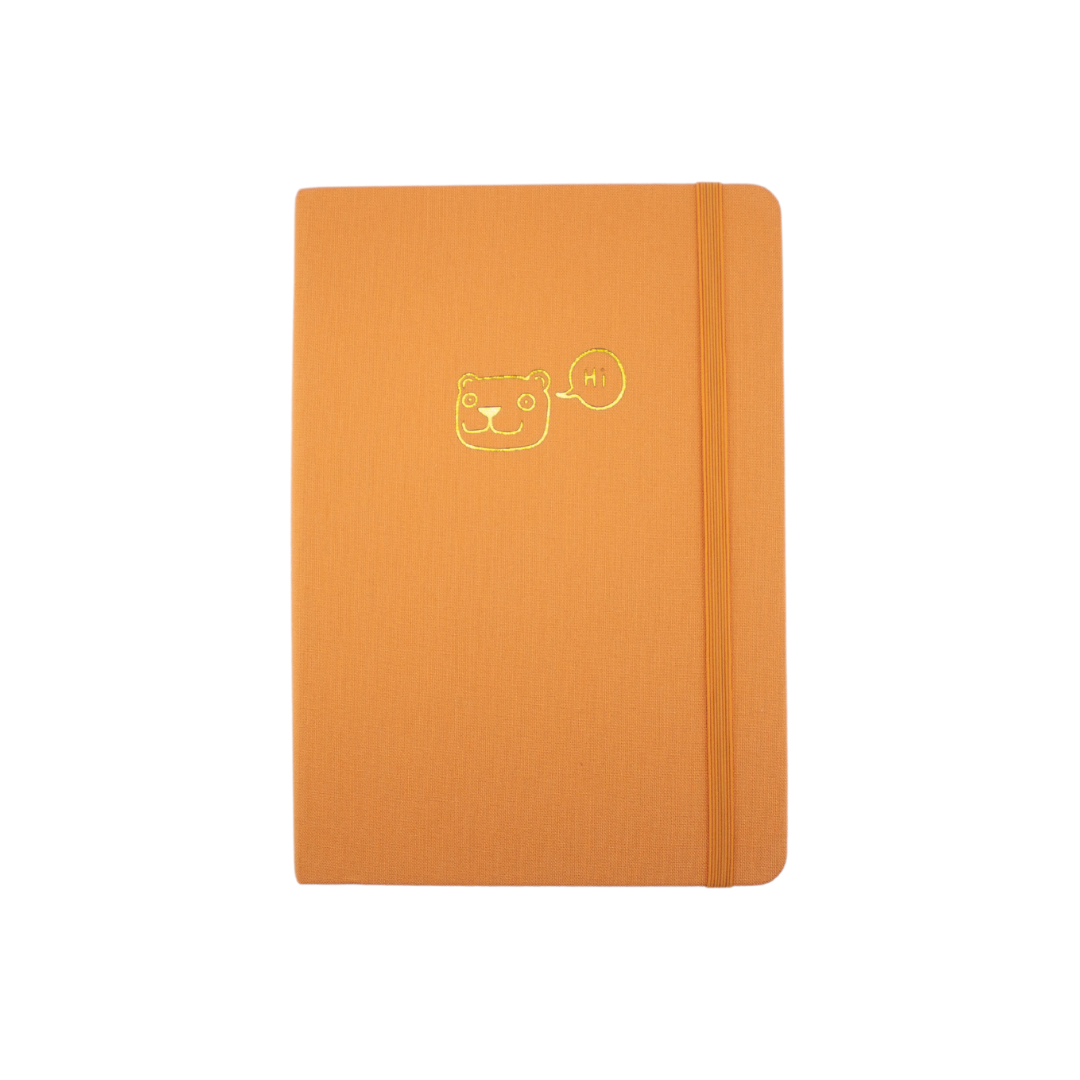The A5 bobo Lined Journal "Tiger" with a tangerine orange linen cover. A gold foil smiling cartoon tiger head with a speech bubble with "Hi" inside, is stamped onto the center of the front cover. A corresponding orange elastic is holding the book closed.