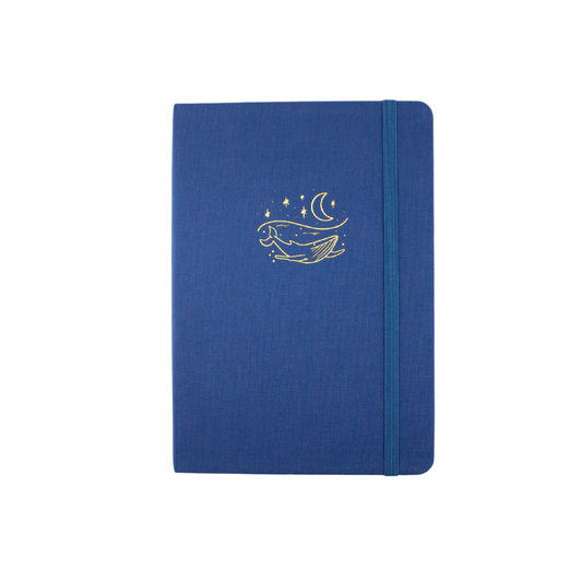 The A5 bobo Lined Journal "Whale" with a deep blue linen cover. A gold foil illustration of a humpback whale in the water under a starry night sky is stamped on the cover. A corresponding medium-dark blue elastic is holding the journal closed.