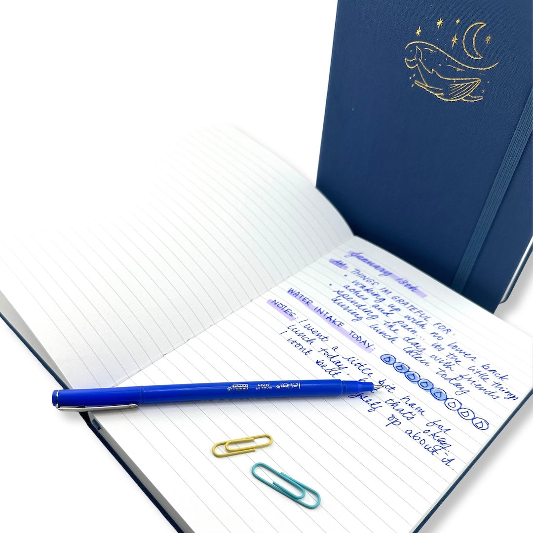 open bobo lined journal with writing on one page and a blue le pen on top with two paper clips. a closed journal with a gold whale and moon decal