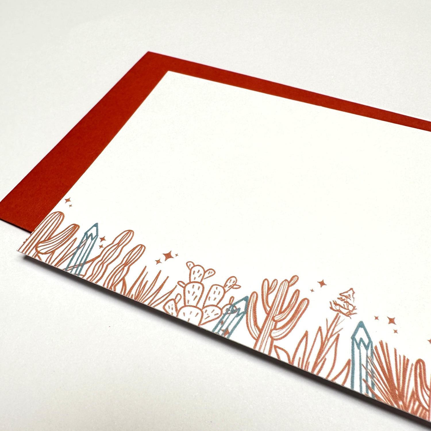 Close up of flat card and rust red envelope on white background. Flat card is 3.5 x 5 inches, landscape orientation, white background with illustrations on the lower fifth of the card. The illustration is a line drawing of cactus, sparkle stars, and agave in rust red ink and four pencils pointy side up in blue ink hiding amongst the plants.