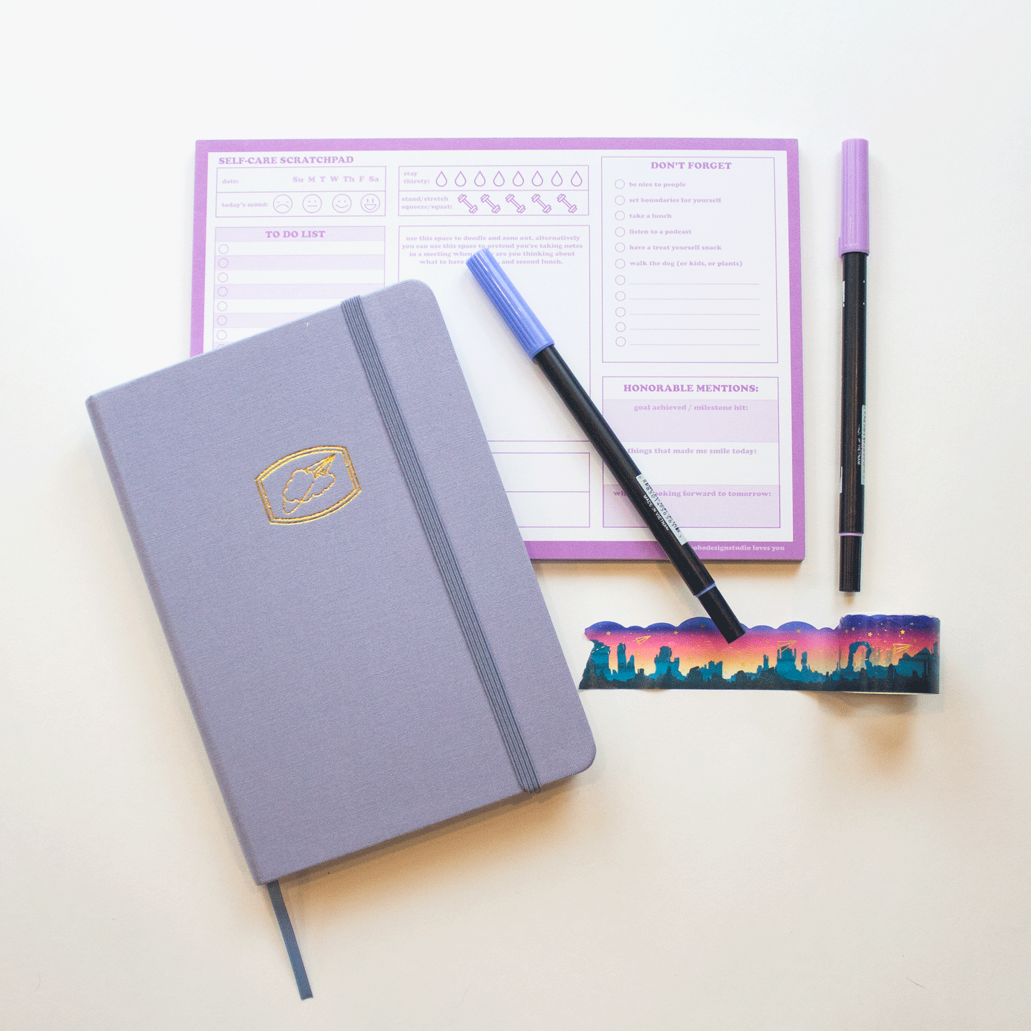 The light blue "Cloud 9" bobo BuJo Dot Grid Journal sits with other desk supplies. The corresponding light blue ribbon bookmark and elastic closure are visible. Markers, bobo Southwest Sunset Washi tape, and a bobo self-care scratchpad are in the background.