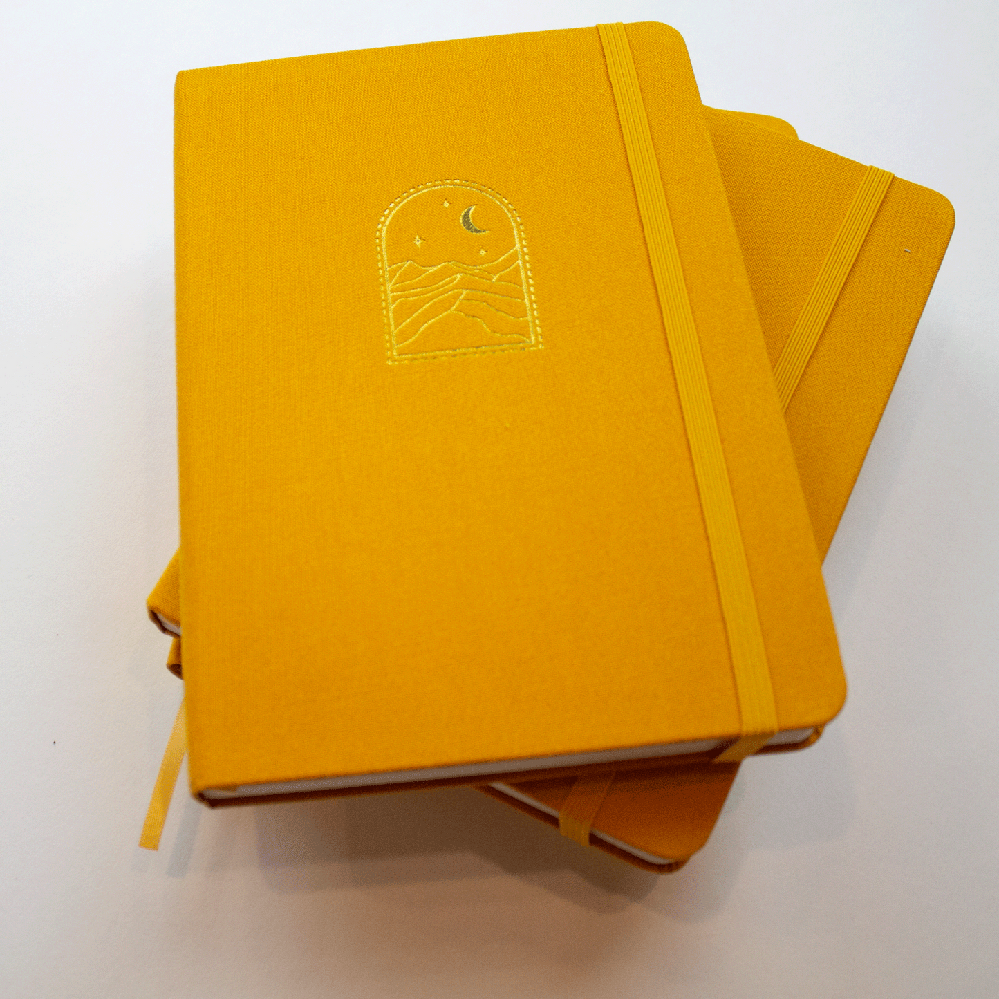A stack of three mustard yellow "Golden Dunes" bobo dot grid journals. The gold stamped starry night sky and sand dune illustration on the front cover can be seen on the first book. Their matching yellow elastic closures and ribbon bookmarks can also be seen. 