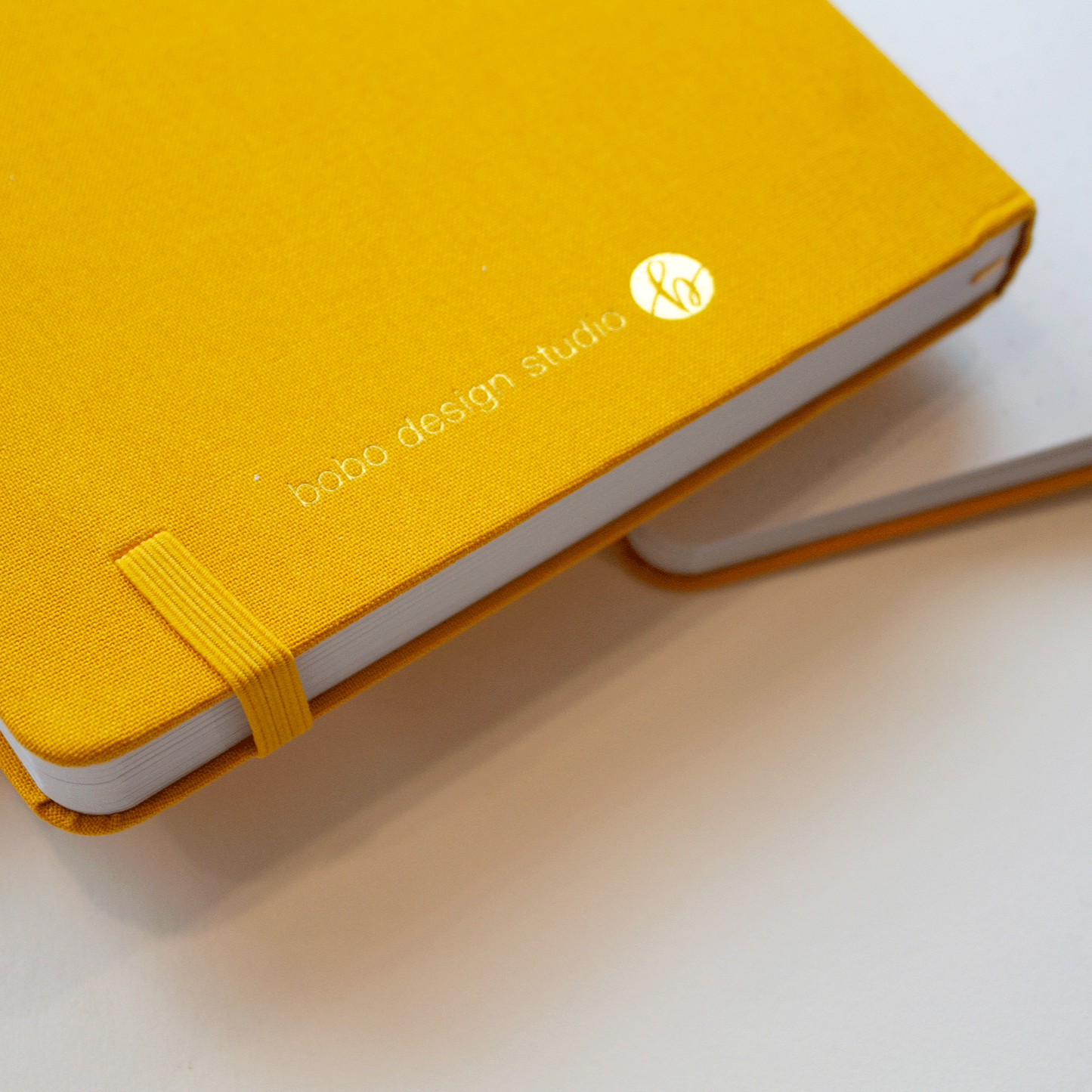 Two Golden Dunes bobo BuJo Dot Grid Journals. The bottom journal is lying open in the background. The journal on top is showing its back cover with the gold stamped text "bobo design studio" and logo of a cursive lowercase b in a circle. The end point where the yellow elastic closure fits into the yellow linen cover is visible.