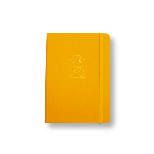 The A5 "Golden Dunes" bobo BuJo dot grid journal with mustard yellow linen cover. The matching yellow elastic closure holds the journal closed. A gold foil illustration of sand dunes under a starry night sky and crescent moon is stamped onto the front cover.