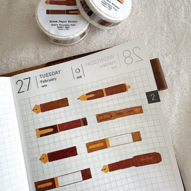 Two small round containers of Bloom Paper Studio washi tape rolls placed on a light fabric surface, with an open planner showing illustrated washi tape designs spread across the page.