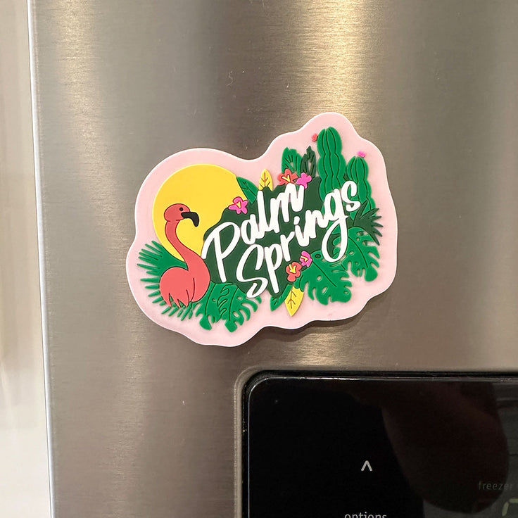 Palm Springs flamingo magnet. features a flamingo with a sun behind it and leaves with some cactus and "palm springs" written in white cursive letters with a pink background on a fridge 