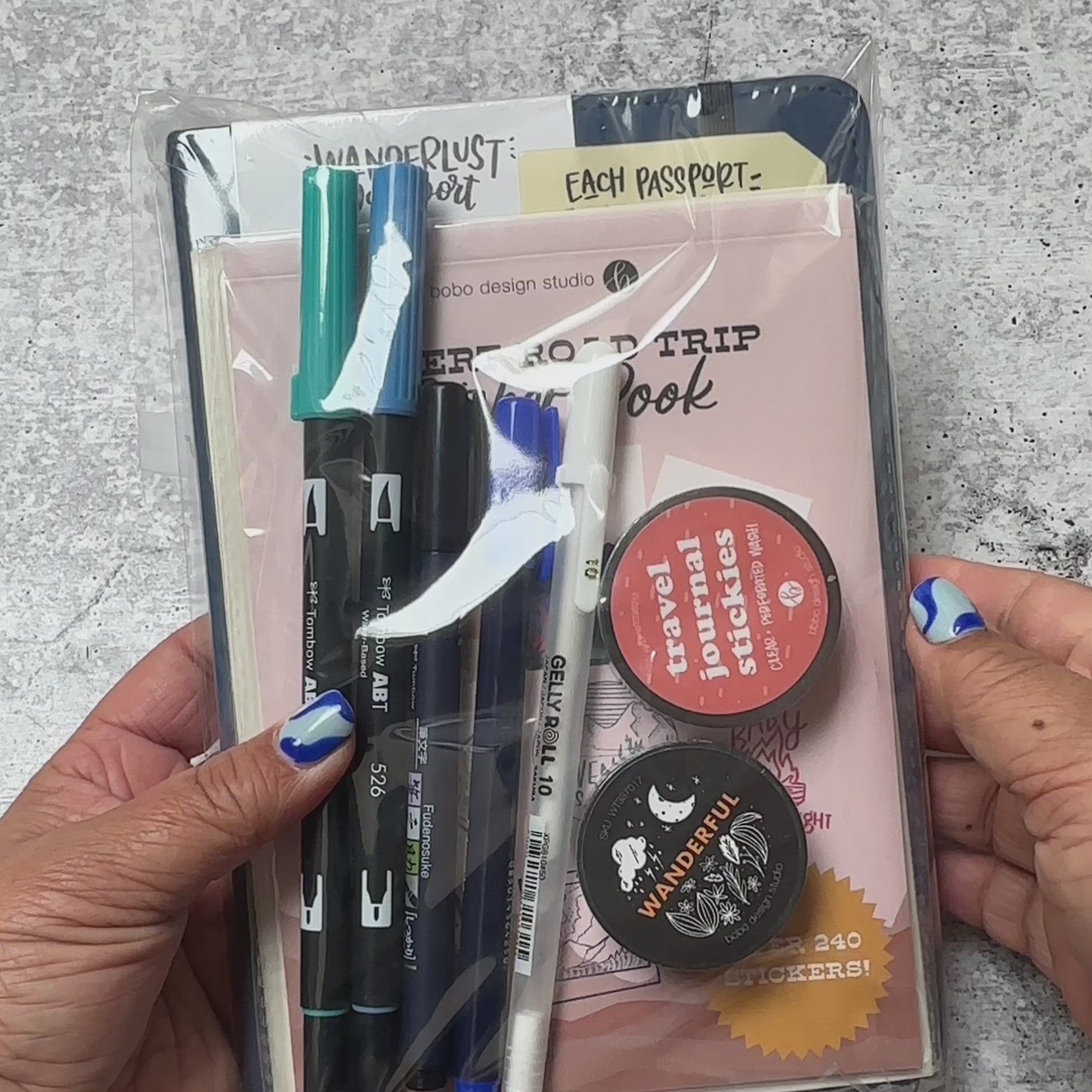 a video that showcases what comes in a wanderlust passport bundle. A navy blue wanderlust passport with a sticker book, two sticker sheets, a sticker book, a black wanderful washi tape, a clear washi tape with words, two blue tombow duel brush pens, a black and a blue tombow fudenosuke brush pen, and a white gelly roll gel pen