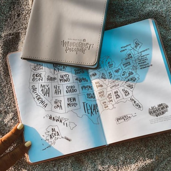 Two Wanderlust Travel Journals lying on the beach. One is opened to the color-in US Map Travel Tracker, with includes 50 US states and Puerto Rico