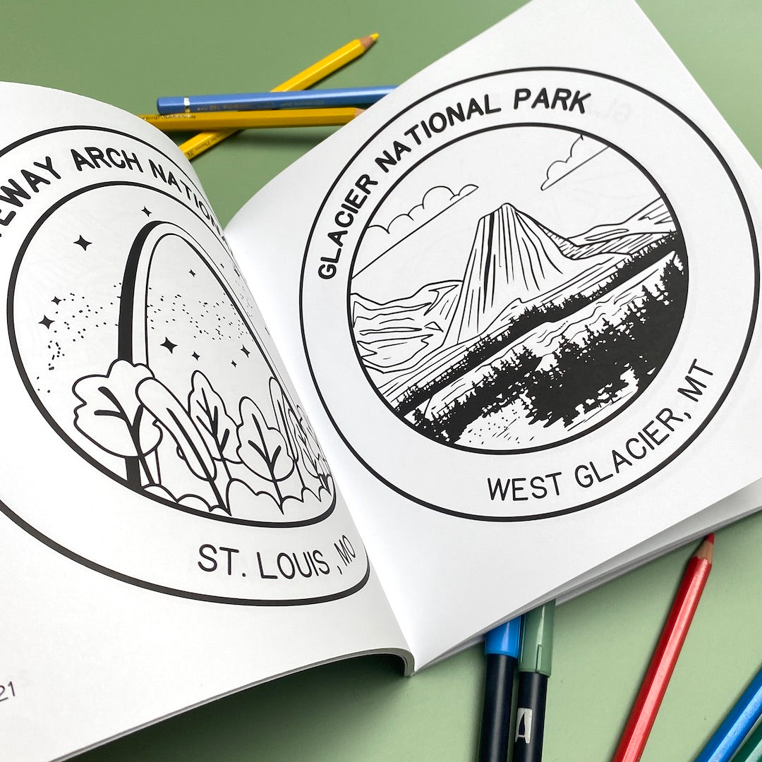 The National Parks Coloring Book is opened to pages 21 and 22. Page 21 has a black line drawing of Gateway Arch National Park, MO printed on the white page. Page 22 is printed with a line drawing of Glacier National Park, MT. Coloring pencils and markers are next to the book.