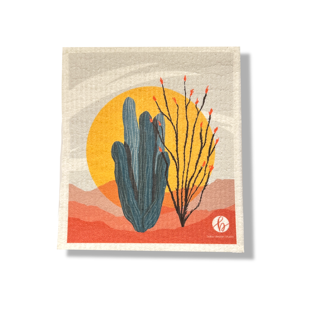 Supercloth on a white background. The sponge cloth is printed with an abstract desert scene, with an organ pipe cactus and ocotillo with red flowers silhouetted on a yellow sun, and growing from red hills and mountains. A small bobo design studio logo is in the bottom right corner, and is a hand lettered cursive "b" in a circle.