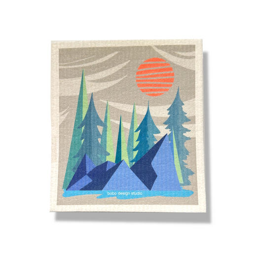Portrait orientation sponge cloth (6.5x8 inches) with vertical wrinkled spongy texture. Printed image centered on cloth with white border. Image of grey sky with abstract white clouds, orange sun, green trees. Dark blue geometric rocks and an aqua blue pond in the foreground.
