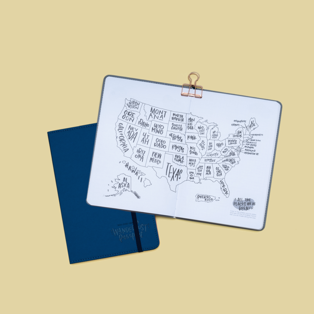 Wanderlust Passport Travel Journal A5 Notebook in Navy Blue with elastic closure, a second example book is open to the US color-in state travel map page spread