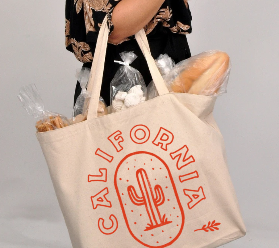An arm holds the California tote bag. Inside the tote bag is a farmers market haul, including a baguette, meringues, and other pastries. An image of a saguaro cactus under the word California is printed on the side in orange.
