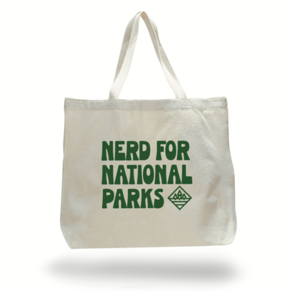 Large natural canvas colored tote bag with "NERD FOR NATIONAL PARKS" printed in green on the side. A small simple image of 3 trees in a diamond shape is printed beside the word PARKS.
