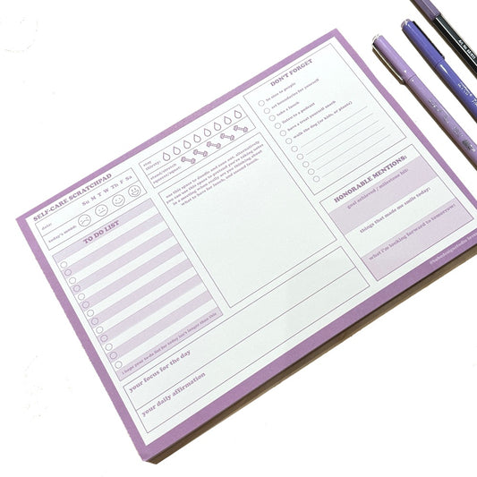 Lavender Self-Care Scratchpad on a white surface, with purple pens beside it. Scratchpad is 10 inch wide, 7 inch tall, day-by-day planning notepad. Three columns of trackers and note-taking prompts, including: date, todays mood, to do list, daily focus, daily affirmation, hydration, exercise, don't forget, goals, and made me smile.