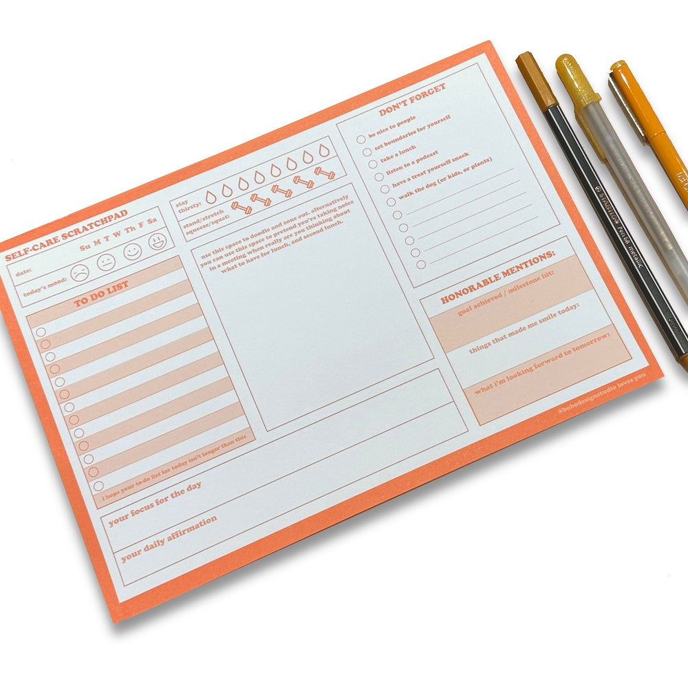 Orange Self-Care Scratchpad on a white surface with orange pens beside it. Scratchpad is printed in landscape orientation (10 inches wide, 7 inches tall). Three columns of daily planning trackers and notetaking boxes are visible, including: date, today’s mood, to-do list, daily focus, daily affirmation, hydration tracker, exercise tracker, don’t forget list, goals box, and made me smile box. 
